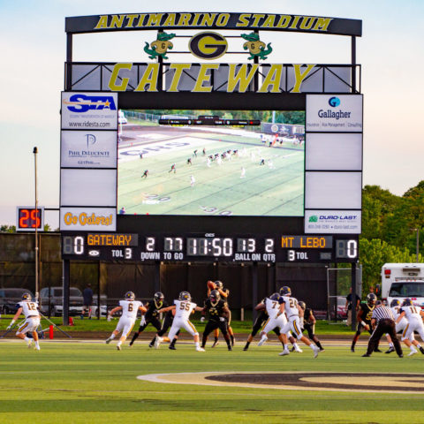 Outdoor scoreboard while football is being played in front.