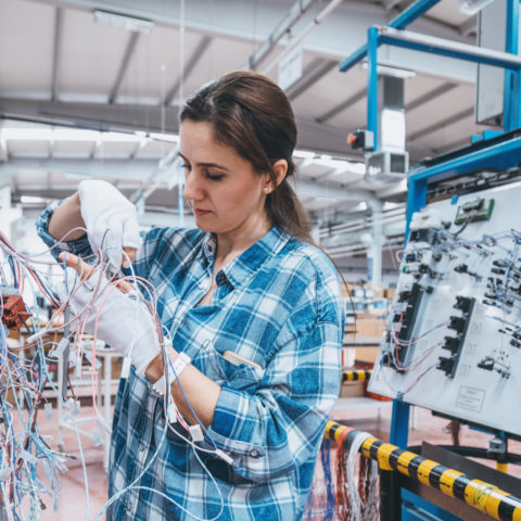 Young women working on wires with a large circuit in the background.