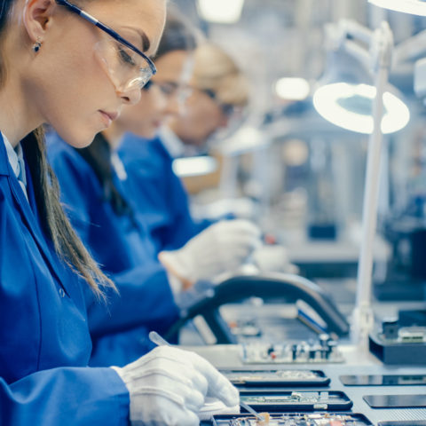 women in blue lab coat working on circuit board with other women also working in background.
