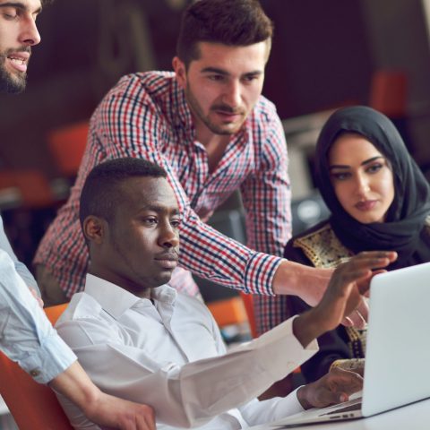 Man pointing at computer screen while team members stand around also looking at screen. 